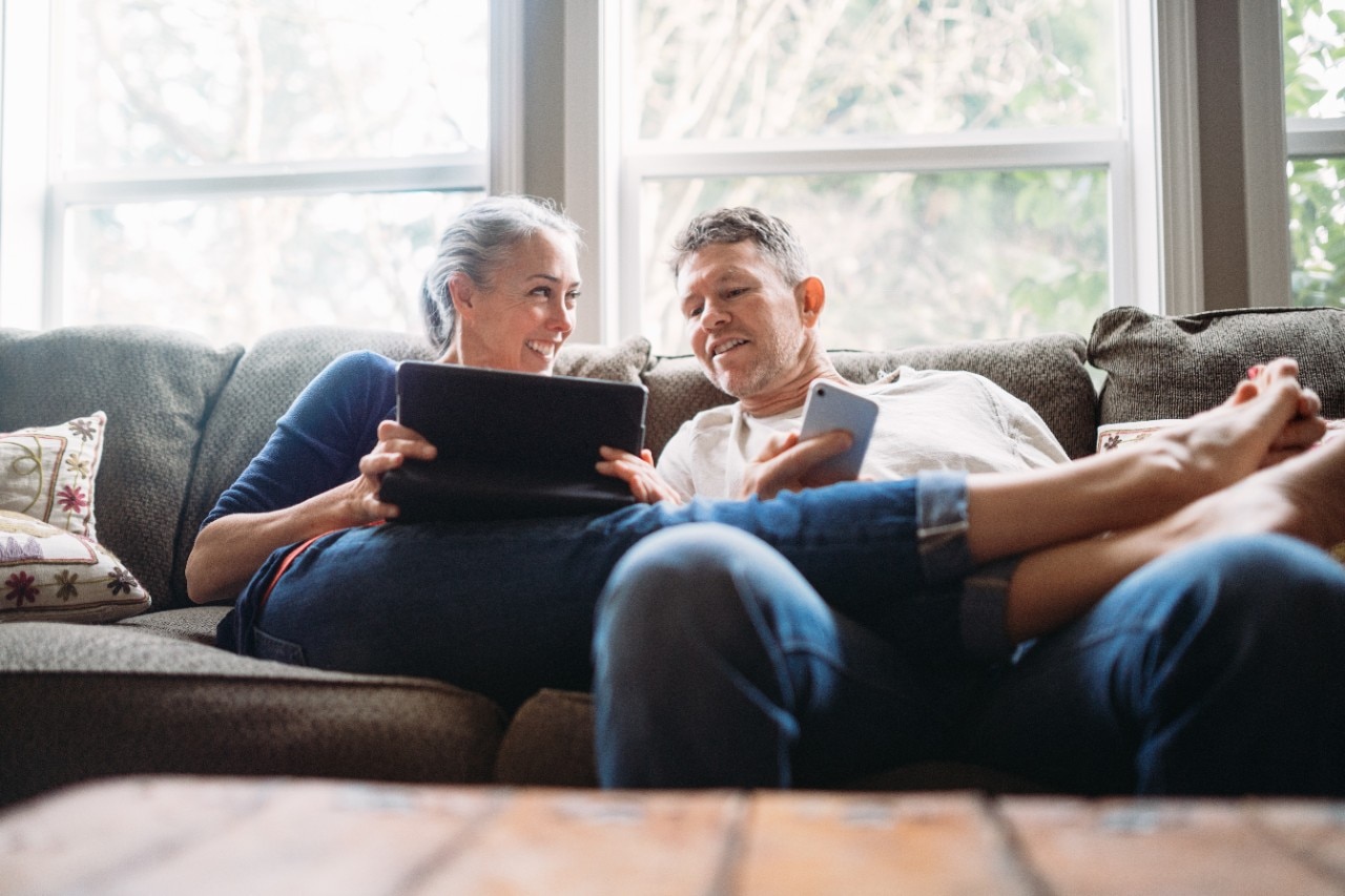 A couple in their 50's relax in their home on the living room couch, enjoying reading and surfing the internet on their mobile touchscreen phones and computer tablet.